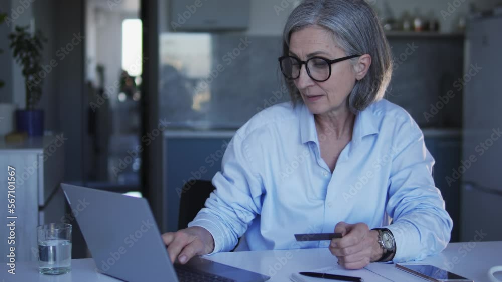 Mature woman in big eyeglasses doing shopping online, entering credit card number into web-site on laptop, choosing goods and paying for home delivery Mature woman in big eyeglasses doing shopping online, entering credit card number into web-site on laptop, choosing goods and paying for home delivery