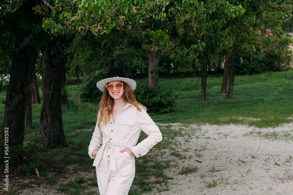 A girl with blond curly hair is dressed in a light overalls and a hat, in sunglasses, stands on the beach and smiles. Hands in pockets. Around the sand against the backdrop of a green park.