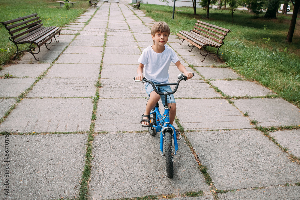 Fototapeta premium A little cute boy in denim shorts and a white t-shirt rides a children's bike along a path in the park during the day.