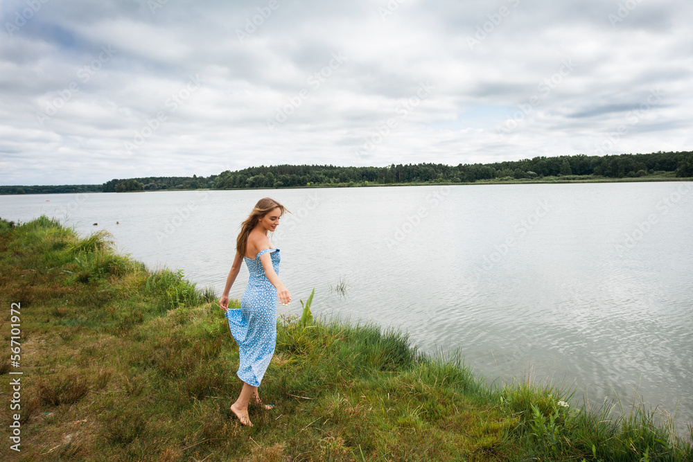 A beautiful slender smiling girl with long blond hair, in a blue summer dress, spins and walks along the river bank, against the backdrop of a picturesque landscape. A beautiful slender smiling girl with long blond hair, in a blue summer dress, spins and walks along the river bank, against the backdrop of a picturesque landscape.