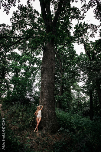 A slender beautiful girl with a sports figure, in light shorts and a T-shirt, stands barefoot near a tall tree in the forest.