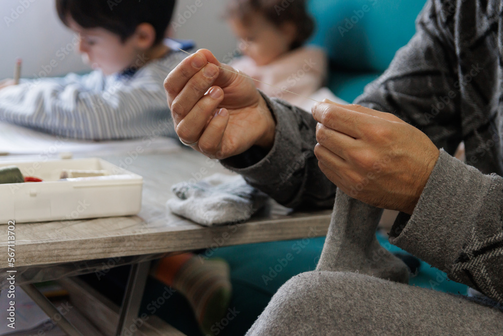 Fototapeta premium Man sewing a sock in the living room at home while his children do their homework