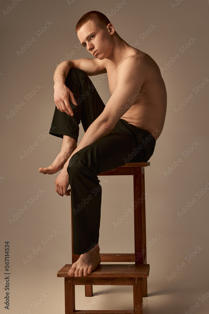Serious. Portrait of handsome young boy with short red hair posing on chair shirtless in pants over light brown studio background. Concept of men's health and beauty, body care, fitness. Body art Serious. Portrait of handsome young boy with short red hair posing on chair shirtless in pants over light brown studio background. Concept of men's health and beauty, body care, fitness. Body art