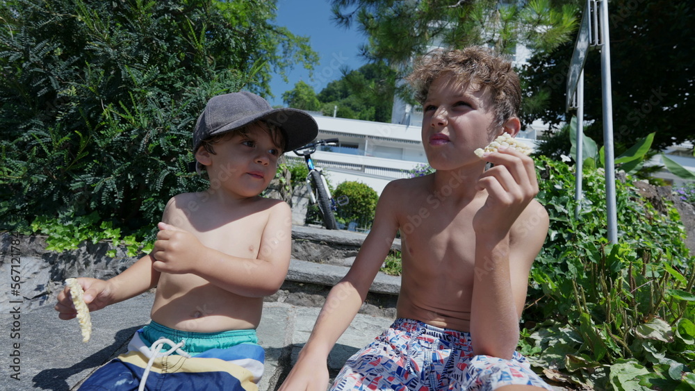Two little brothers hanging out together outside. Younger brother nex to older sibling during summer holidays in the sunlight shirtless. Kids friends eating snack
