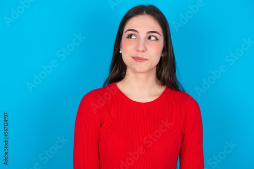 young brunette girl wearing red T-shirt against blue wall looking aside into empty space thoughtful