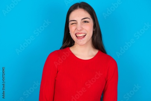 Coquettish young brunette girl wearing red T-shirt against blue wall smiling happily, blinking at camera in a playful manner, flirting with you.