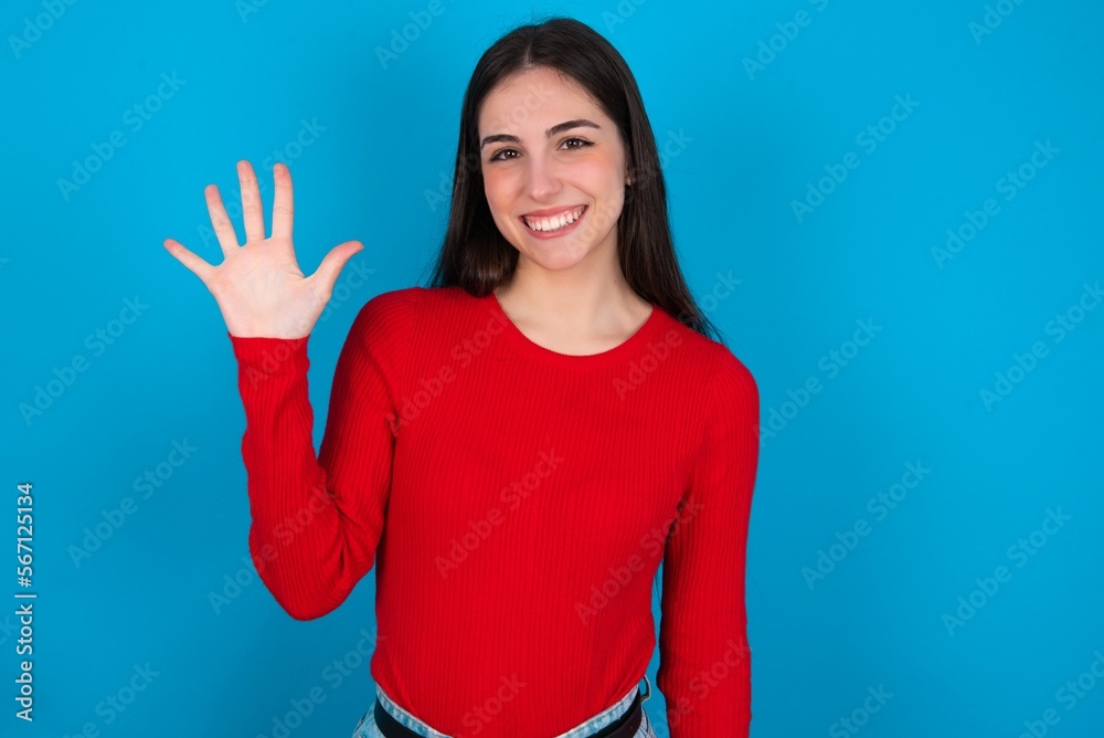 young brunette girl wearing red T-shirt against blue wall showing and pointing up with fingers number five while smiling confident and happy.