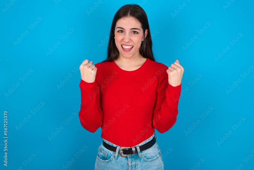 young brunette girl wearing red T-shirt against blue wall celebrating surprised and amazed for success with arms raised and open eyes. Winner concept. young brunette girl wearing red T-shirt against blue wall celebrating surprised and amazed for success with arms raised and open eyes. Winner concept.