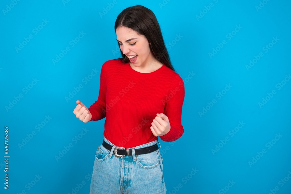 young brunette girl wearing red T-shirt against blue wall very happy and excited doing winner gesture with arms raised, smiling and screaming for success. Celebration concept. young brunette girl wearing red T-shirt against blue wall very happy and excited doing winner gesture with arms raised, smiling and screaming for success. Celebration concept.