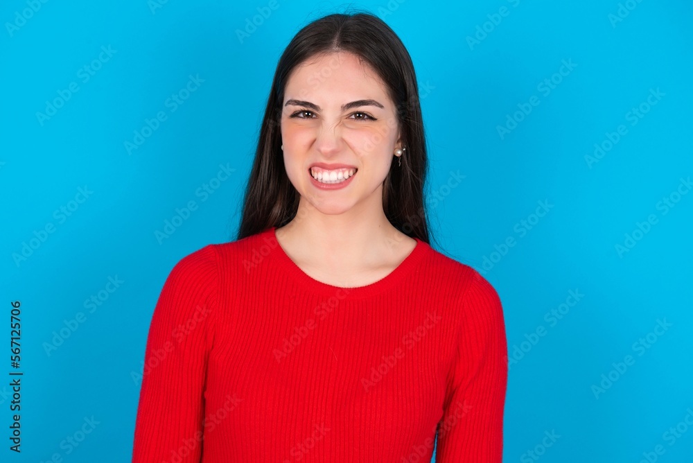 young brunette girl wearing red T-shirt against blue wall keeps teeth clenched, frowns face in dissatisfaction, irritated because of much duties. young brunette girl wearing red T-shirt against blue wall keeps teeth clenched, frowns face in dissatisfaction, irritated because of much duties.