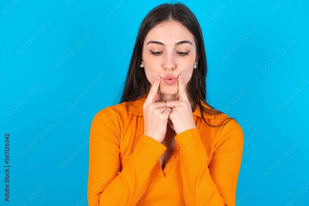 young caucasian brunette girl wearing orange crop top against blue wall crosses eyes and makes fish lips funny grimace young caucasian brunette girl wearing orange crop top against blue wall crosses eyes and makes fish lips funny grimace