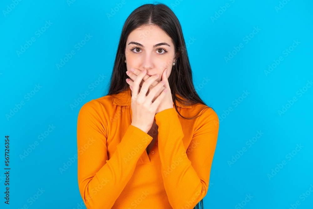 Stunned young caucasian brunette girl wearing orange crop top against blue wall covers both hands on mouth, afraids of something astonishing