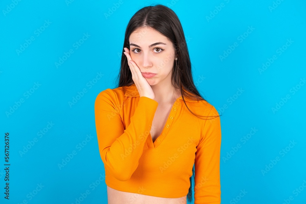 Sad lonely young caucasian brunette girl wearing orange crop top against blue wall touches cheek with hand bites lower lip and gazes with displeasure. Bad emotions Sad lonely young caucasian brunette girl wearing orange crop top against blue wall touches cheek with hand bites lower lip and gazes with displeasure. Bad emotions