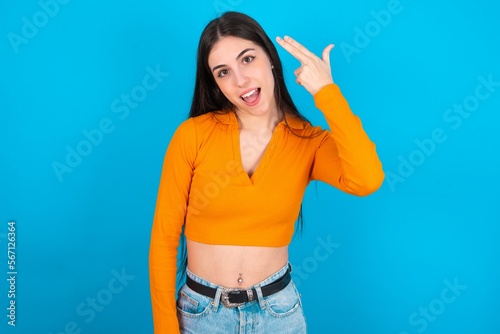 Obraz na plátně young caucasian brunette girl wearing orange crop top against blue wall foolishness around shoots in temple with fingers makes suicide gesture