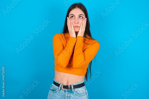 young caucasian brunette girl wearing orange crop top against blue wall keeps hands on cheeks has bored displeased expression. Stressed hopeless model