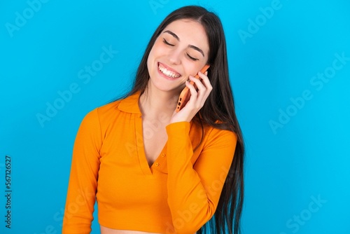 Funny young caucasian brunette girl wearing orange crop top against blue wall laughs happily, has phone conversation, being amused by friend, closes eyes.