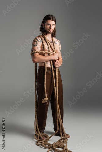 Full length of tattooed man with rope on body on grey background.