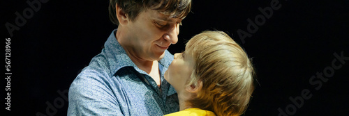 man father dad holds little kid boy son six years old in his arms and hugs. happy family, parenthood, fatherhood and childhood in studio illuminated by bright light and black background. banner