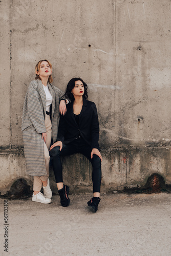 Two pretty women friends posing near gray wall. Couple of gay lesbian girls hugging embracing together girlfriends, dressed casual outfits, have a date. LGBT concept. Fashion, make up, hairstyle 