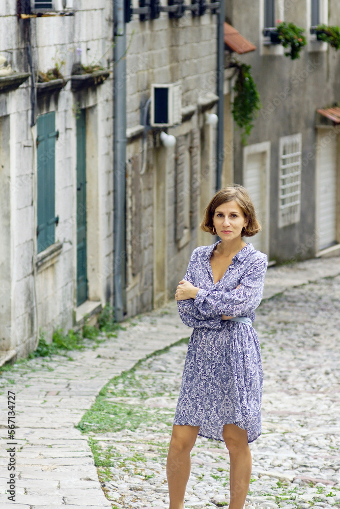 A young woman in a dress stands with folded hands on a cobbled street