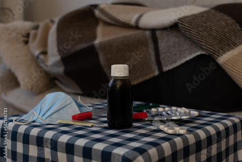 Disposable medical mask, thermometer, potions and pills on blurred background of person under the plaid on sofa