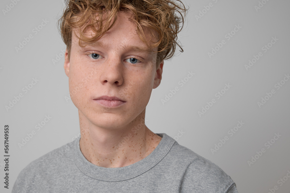 Portrait of young serious handsome freckled red-haired curly boy wearing gray t-shirt and posing for the camera in studio on a gray background. Portrait of young serious handsome freckled red-haired curly boy wearing gray t-shirt and posing for the camera in studio on a gray background.