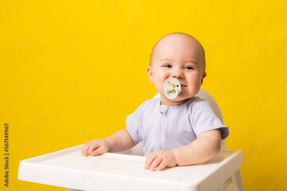 cute baby with a pacifier in his mouth in a high chair on a yellow background. Baby food, space for text cute baby with a pacifier in his mouth in a high chair on a yellow background. Baby food, space for text