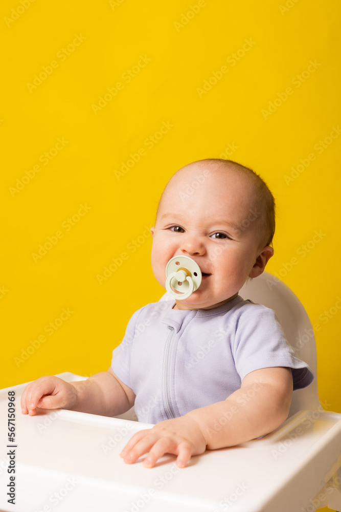 cute baby with a pacifier in his mouth in a high chair on a yellow background. Baby food, space for text cute baby with a pacifier in his mouth in a high chair on a yellow background. Baby food, space for text