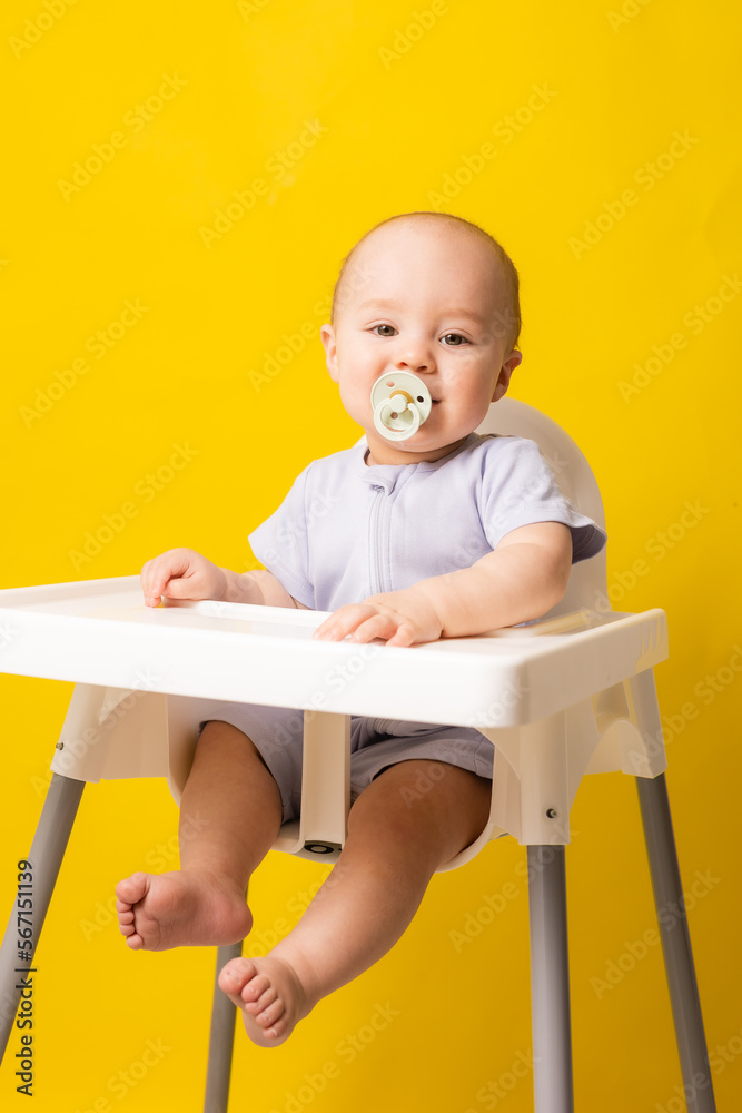 cute baby with a pacifier in his mouth in a high chair on a yellow background. Baby food, space for text cute baby with a pacifier in his mouth in a high chair on a yellow background. Baby food, space for text