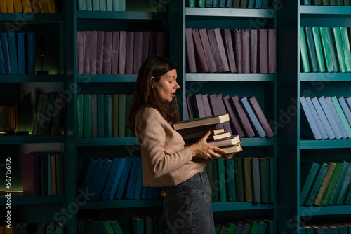 A student in the library, preparing for exams, studying at the university. A young woman holds a stack of heavy textbooks in her hands