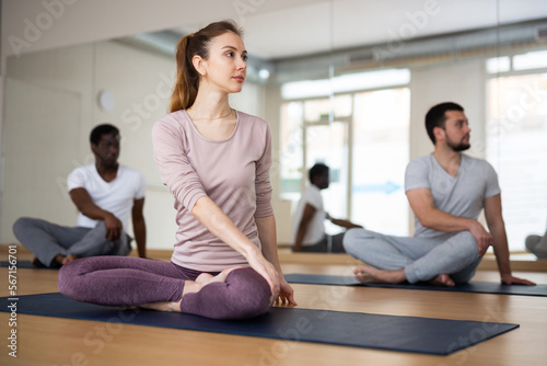 Concentrated young woman performing seated twisting asana Parivrtta Ardha Padmasana during group yoga class in fitness studio..