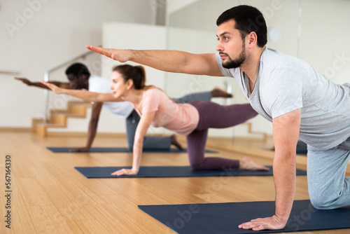 Young bearded man maintaining active lifestyle exercising with group in yoga studio, standing in balancing asana Dandayamna Bharmanasana..