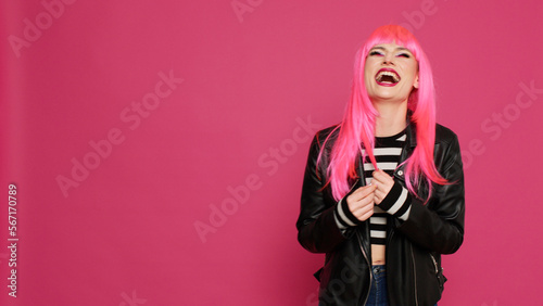 Cheerful lovely woman with pink hair wig laughing and having fun on camera, showing candid smile in studio. Young happy girl with leather jacket enjoying jokes and having laugh.