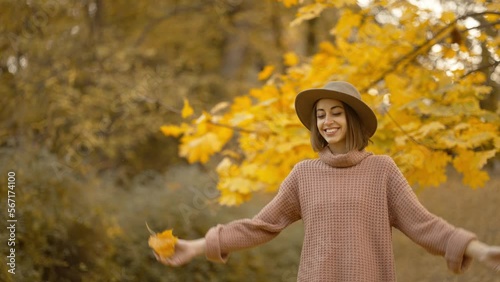 slow motion autumn portrait of attractive smiling woman wearing in brown dress, sweater and hat. girl holds yellow leaves, playfuly looking at camera and throws leaves up photo