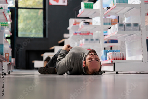 Young caucasian man having epilepsy attack, lying unconscious on floor in medical retail store. Pharmacy store customer fainting, suffering from neurological disease seizure photo
