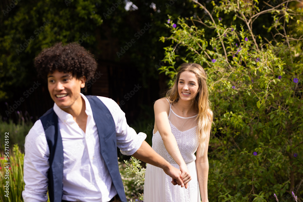 Happy diverse bride and groom holding hands, walking in garden at outdoor wedding Happy diverse bride and groom holding hands, walking in garden at outdoor wedding