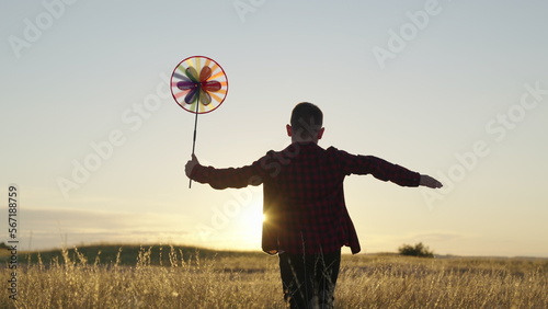 Happy boy playing with toy pinwheel outdoors in summer in park at sunset. Active Kid runs and holds colored wheel on stick in his hand. Boy plays with turntable, autumn field. Family walk in autumn