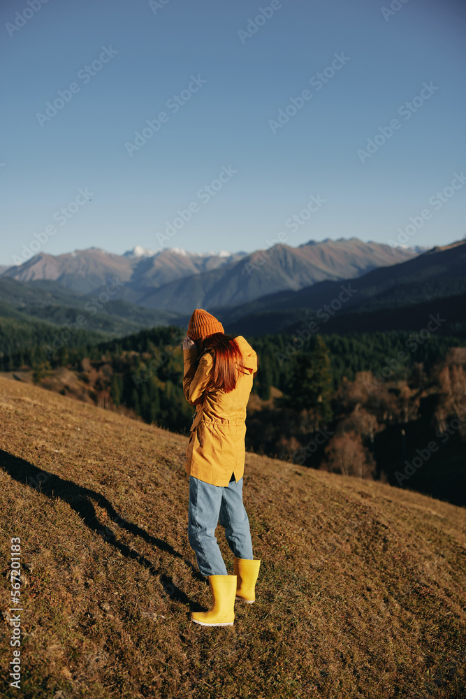 Woman walking full-length on a hill in the autumn and looking at the mountains in a yellow raincoat and jeans happy trip in the sun sunset hiking, freedom lifestyle 