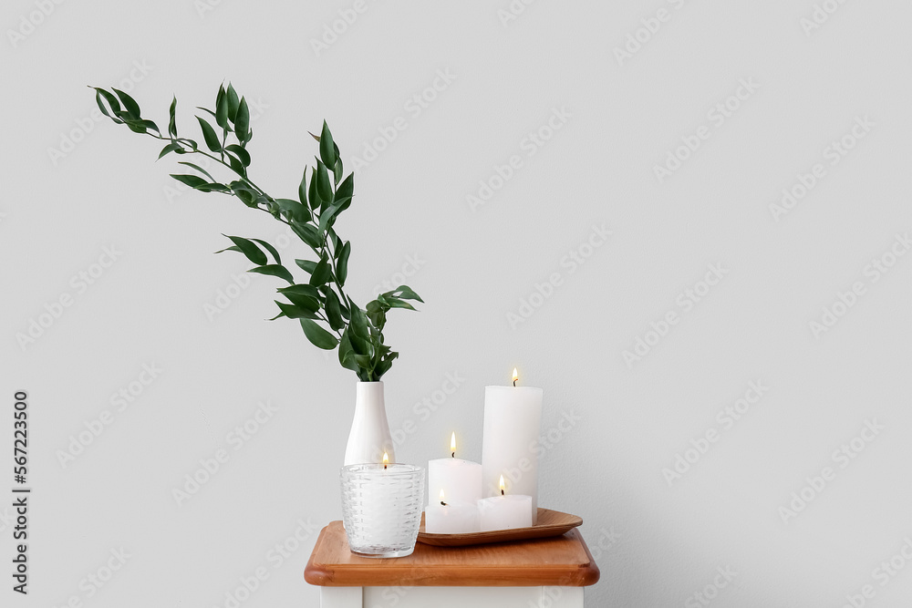 Burning candles and vase with ruscus branches on end table near grey wall Burning candles and vase with ruscus branches on end table near grey wall