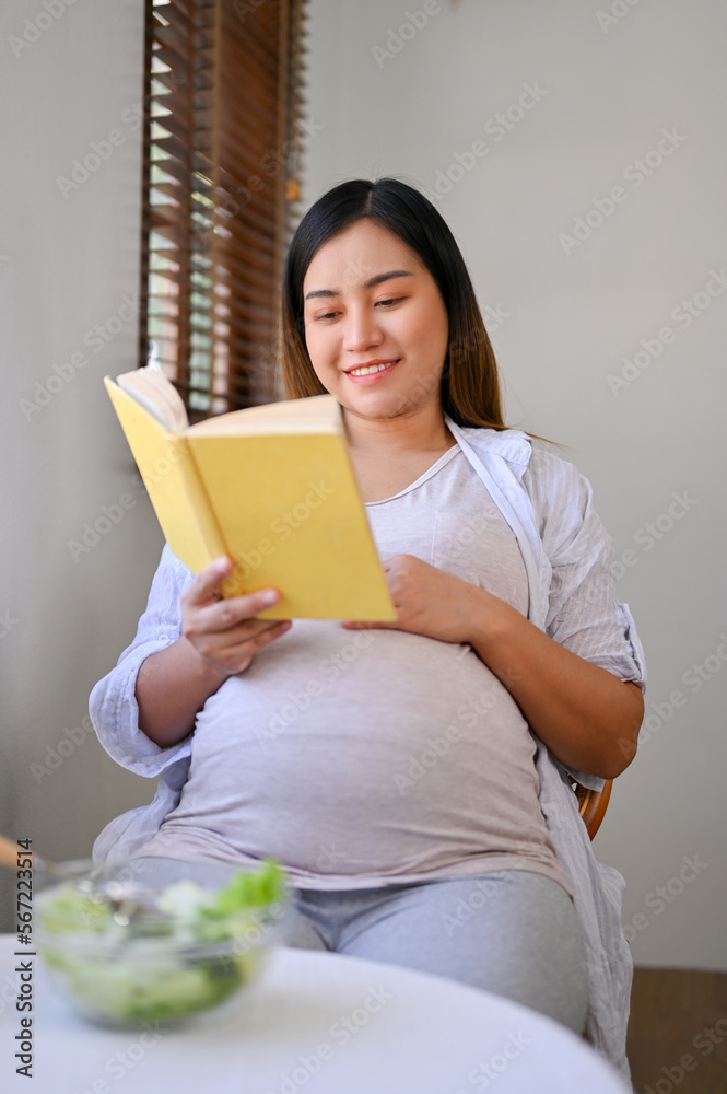 Relaxed Asian pregnant woman reading a book at a dining table, preparing to be a mother. Relaxed Asian pregnant woman reading a book at a dining table, preparing to be a mother.