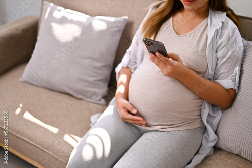 Happy Asian pregnant woman using her phone while relaxing on sofa in her living room.