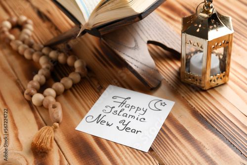 Card with text HAPPY ISLAMIC NEW YEAR, lantern, Koran and prayer beads on wooden background, closeup photo