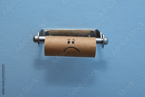 Sad face brown cardboard toilet roll has run out of toilet paper tissues blue wall background middle of frame