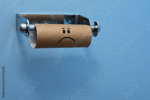 Sad face brown cardboard toilet roll has run out of toilet paper, tissues, blue wall background left of frame
