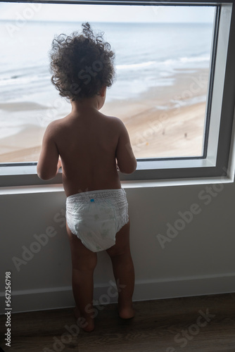 Cute boy toddler on summer vacation looking out of the hotel window at the beach and ocean. He has brown curly hair and is wearing a diaper as seen from back.