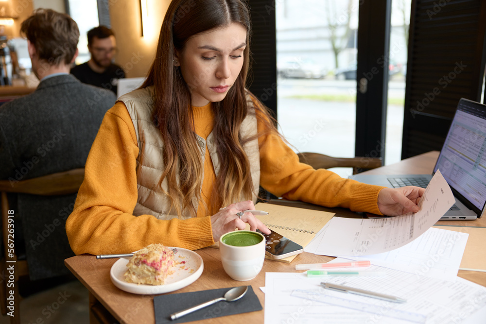 Young caucasian woman taking care of personal finance, sitting at a table in a cafe with bills, laptop and smartphone, working on numbers, using calculator on mobile phone, writing in notebook. Young caucasian woman taking care of personal finance, sitting at a table in a cafe with bills, laptop and smartphone, working on numbers, using calculator on mobile phone, writing in notebook.