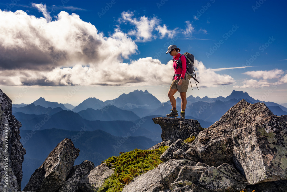 Adventurous athletic female hiker standing on top of a rugged mountain in the Pacific Northwest with jagged mountains in the background.