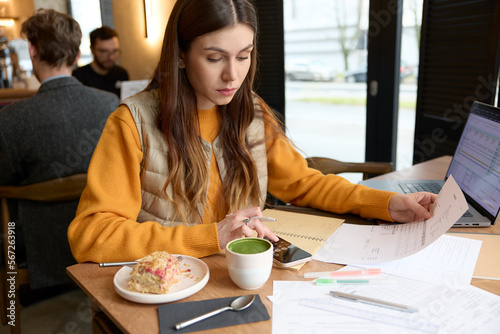 Young caucasian woman taking care of personal finance, sitting at a table in a cafe with bills, laptop and smartphone, working on numbers, using calculator on mobile phone, writing in notebook. 