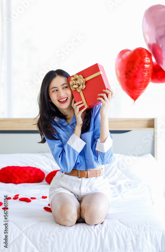 Millennial Asian young romantic female girlfriend sitting on bed in bedroom decorate with pink heart shape balloon holding opening red gold ribbon wrapped surprise gift box on valentine day festive