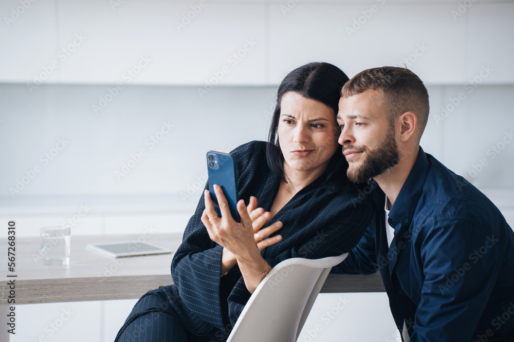Perplexed young couple sitting at kitchen looking at phones screen with puzzled face expression upset by news. Young adult hispanic woman shows to husband, disappointed by economic crisis. Frustration Perplexed young couple sitting at kitchen looking at phones screen with puzzled face expression upset by news. Young adult hispanic woman shows to husband, disappointed by economic crisis. Frustration